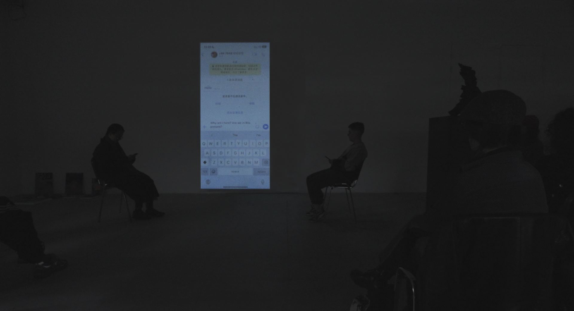 An image of a person sitting in a dark room next to a large, vertical projection of a phone screen with messages on it.