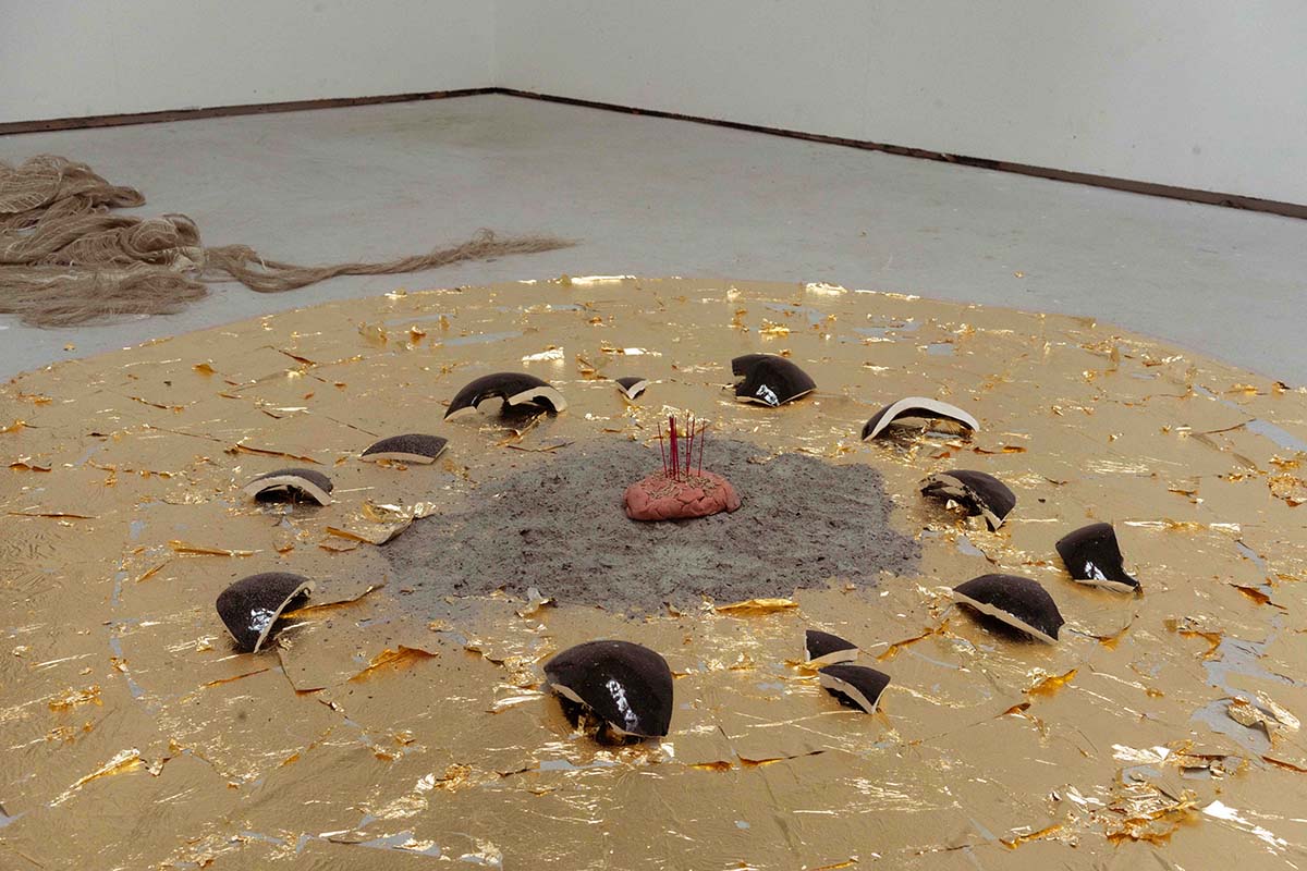 An image of several pieces of broken, black ceramic on gold leaf, arranged in a circle around a mass of red clay holding incense sticks.