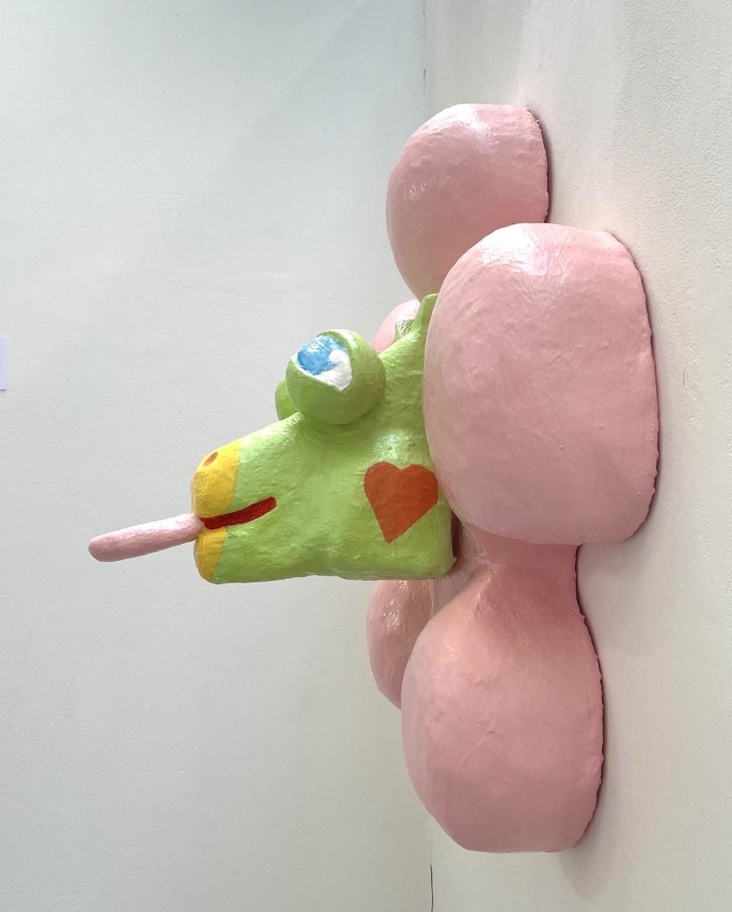 Sculpture on a wall of a green horse with its tongue out mounted onto a pink flower.