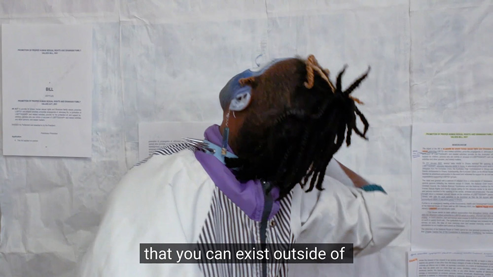 A video still with captioned text. A black person wears blue and white face paint and a blue choker in front of a white background. There are black stripes on their face paint and jacket.