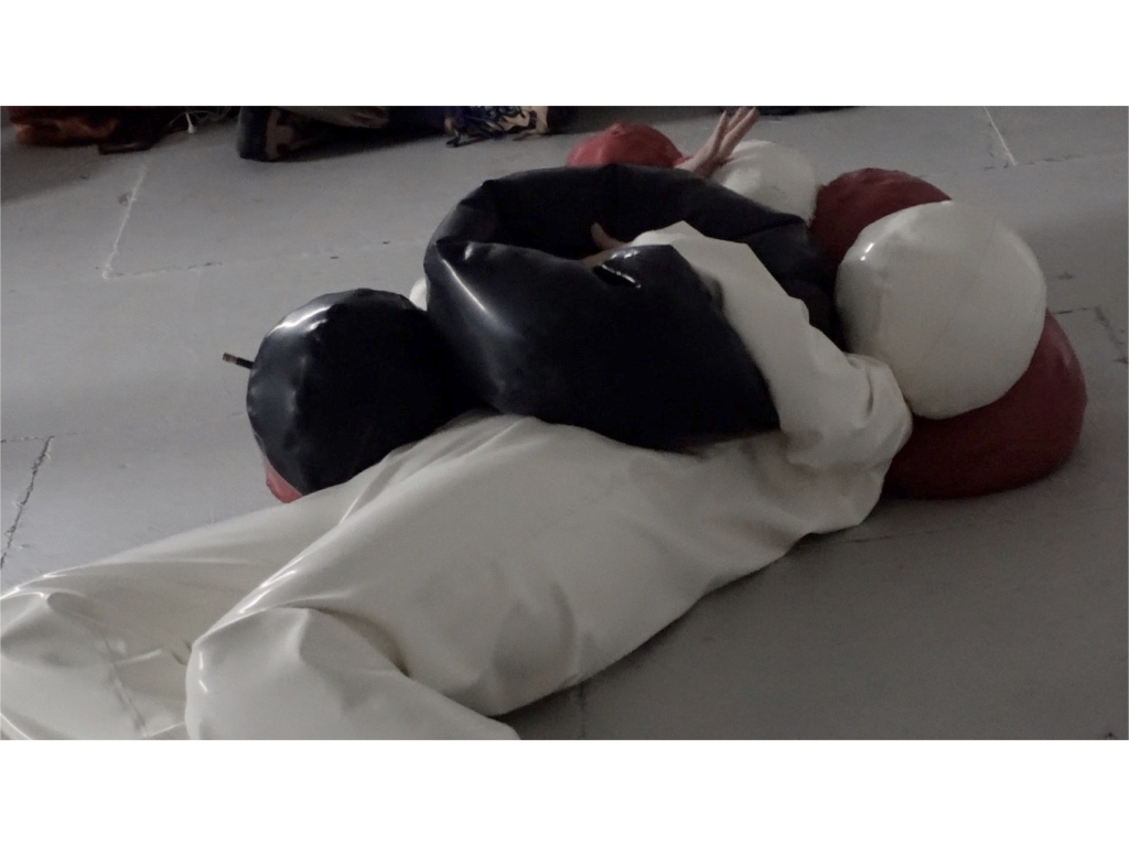 A still from a video where a person is covered in black, red and white balloons.