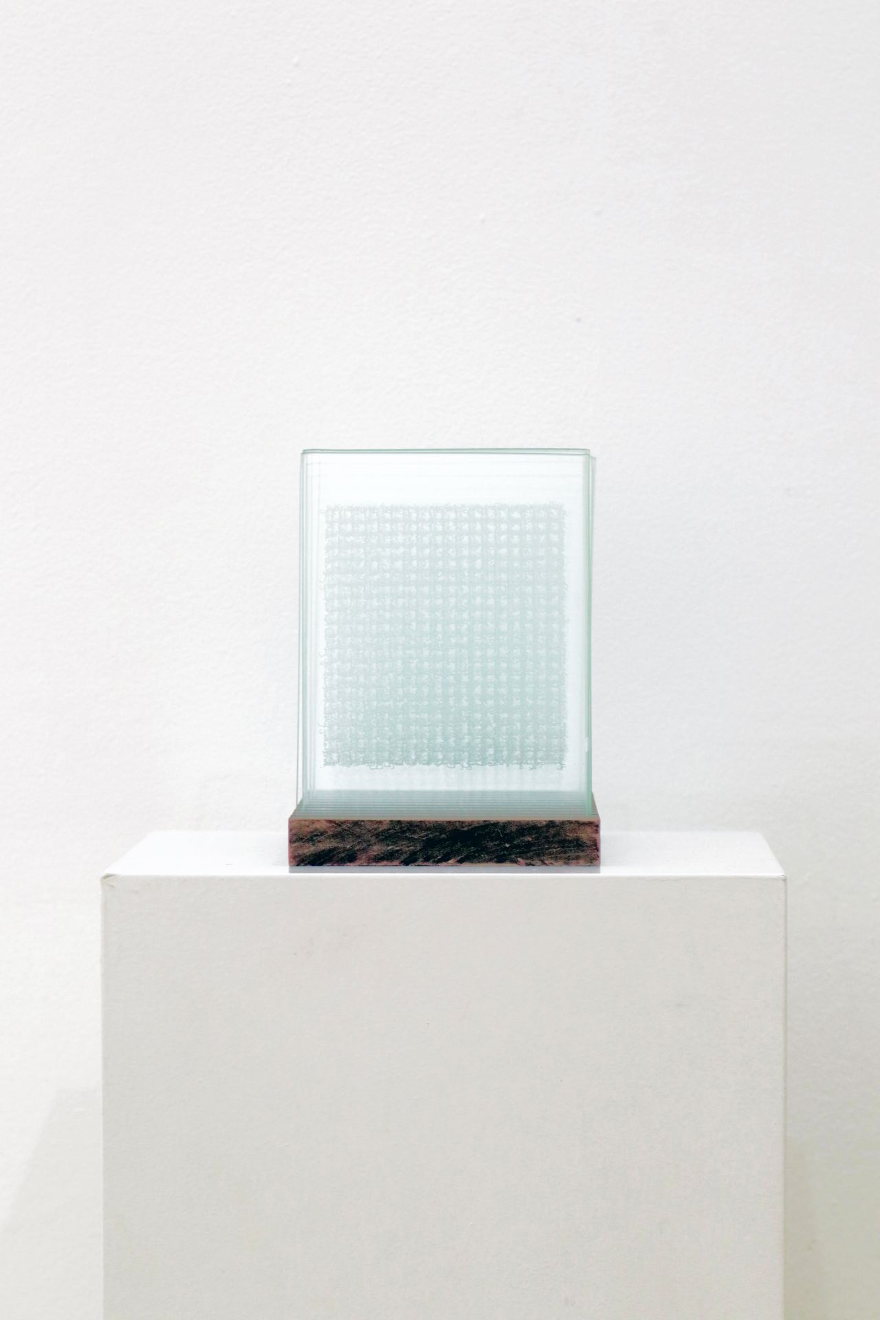 A sculpture made of several rectangular pieces of glass what are etched with writing and set in a wooden block, atop a white plinth.