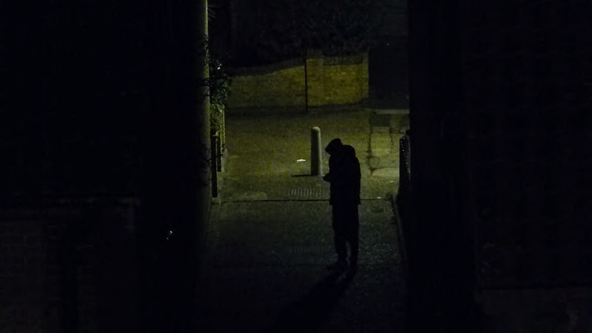 A man stood in an alleyway in an undisclosed location rolls a cigarette while waiting for a phone call. 