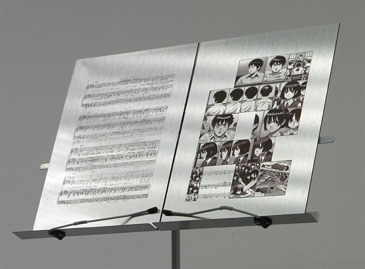 An image of two metal A4 pages on a metal music stand. The page on the left is printed with a score of music. The page on the right is a page from a comic book.