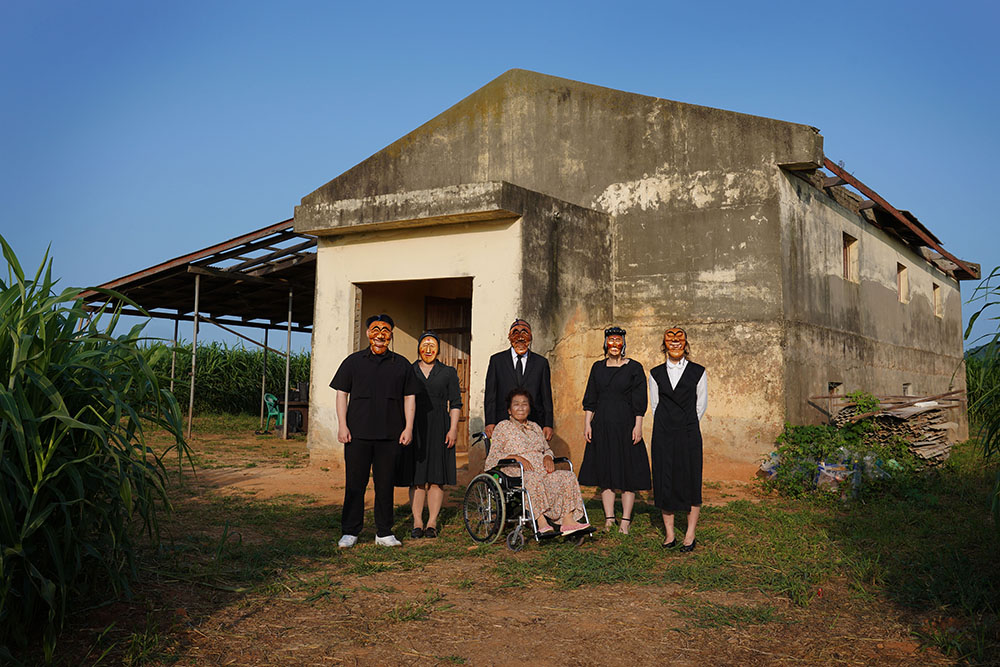 A photograph of a group of people dressed in black, wearing traditional Korean masks that are used to hide emotions, stood in front of a concrete barn set against a blue sky.