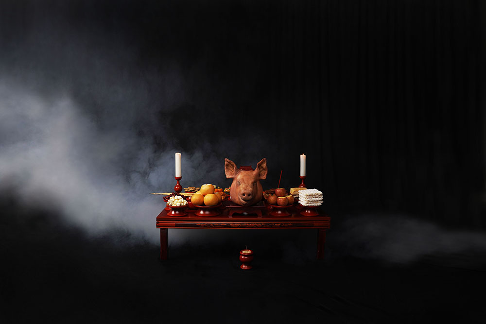 An image of a feast on a wooden table featuring a cooked pig's head and two candlesticks, set against a black, smoky background.