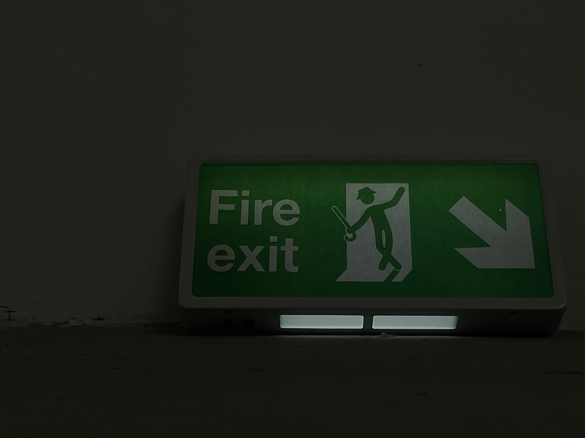 An image of an altered fire-exit sign, showing a figure in a hat and with a walking-stick, dancing.