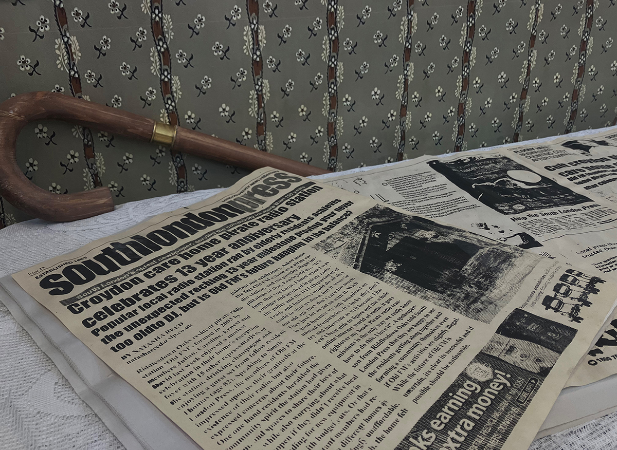 An image of a desk supporting a newspaper and a walking stick, set against a floral-wallpapered wall.