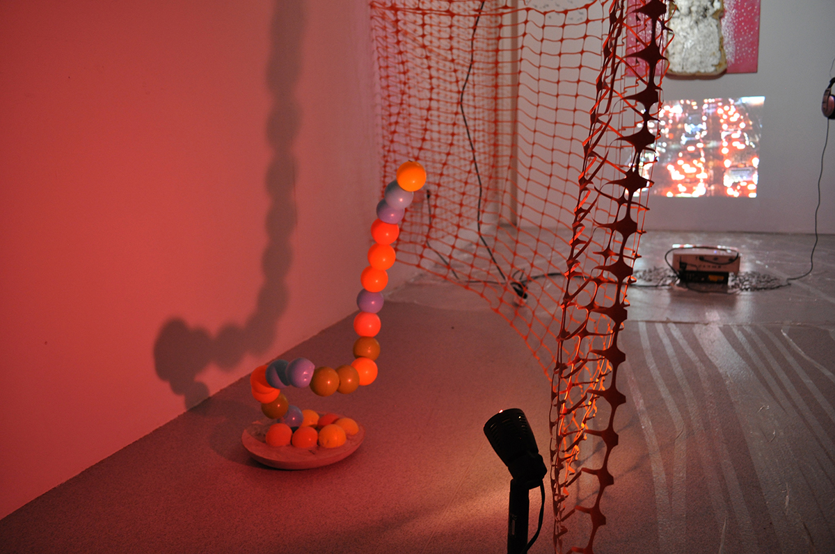 A detail shot of the installation. A sculpture of play-pit balls vertically placed on top of each-other, like a vertically positioned snake. There is a large shadow behind it and the lighting is pink.