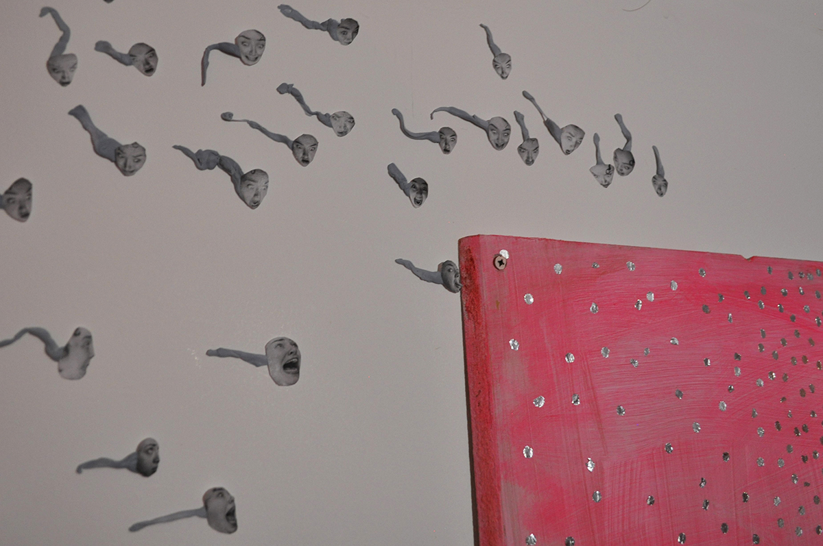 This image shows a corner of the pink painted wooden surface. Blu-tack sperm like shapes gather around its corner. On these shapes are printed out pictures of a women’s face with different facial expressions.