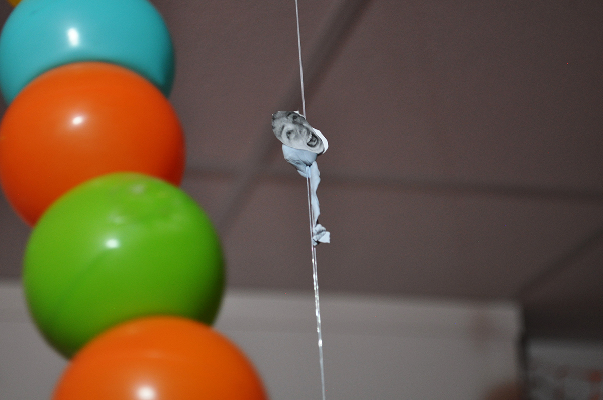 This image is a close-up of the small sperm-shaped blutack on fishing wire with a women’s face printed out on a piece of paper stuck onto one end of it. The background has play-pit balls.