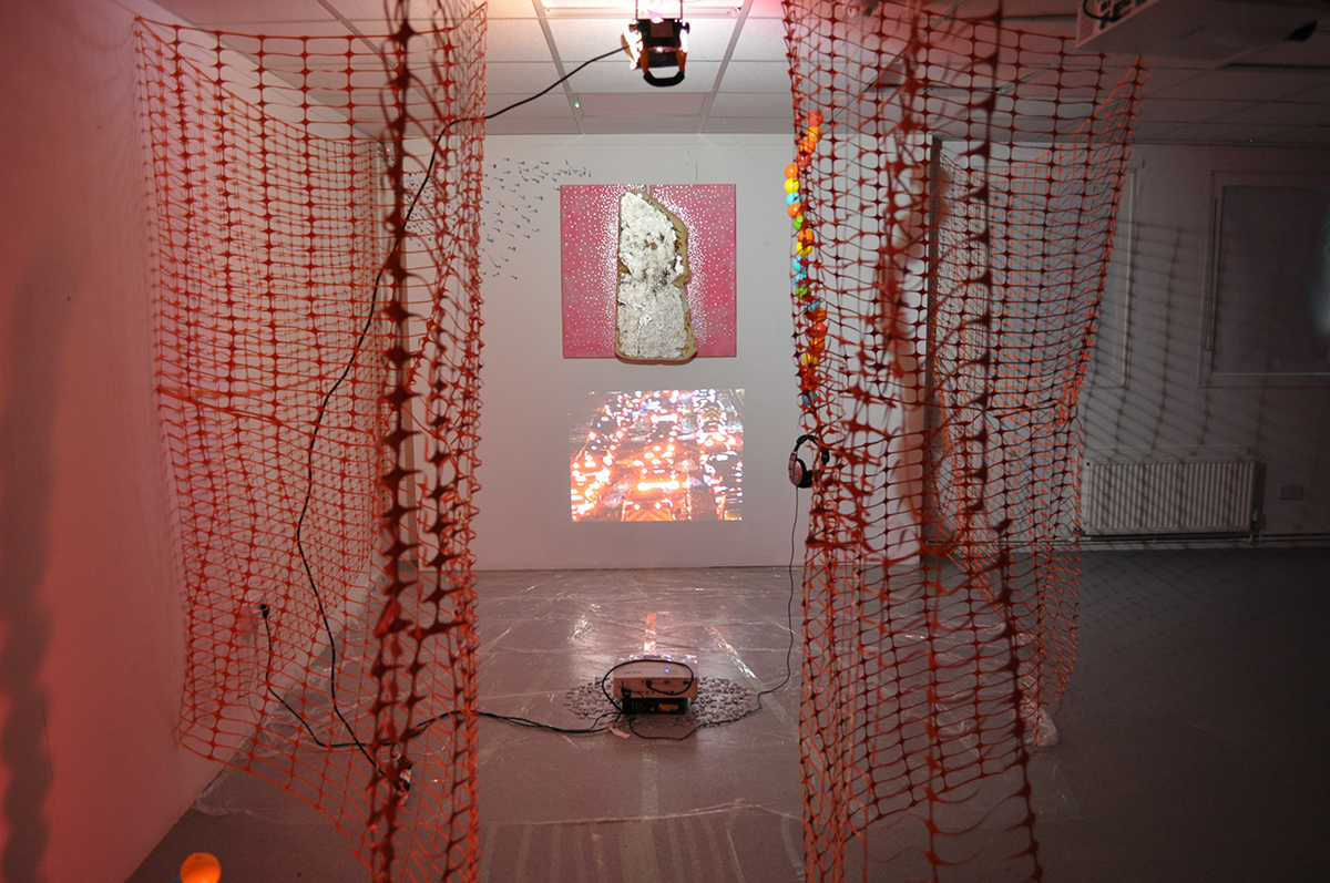 An art installation, containing plastic construction fencing, a projection of a queue of cars, a sculpture of insulation foam on pink painted wood and silver foil dots. Plastic sheeting is on the floor.