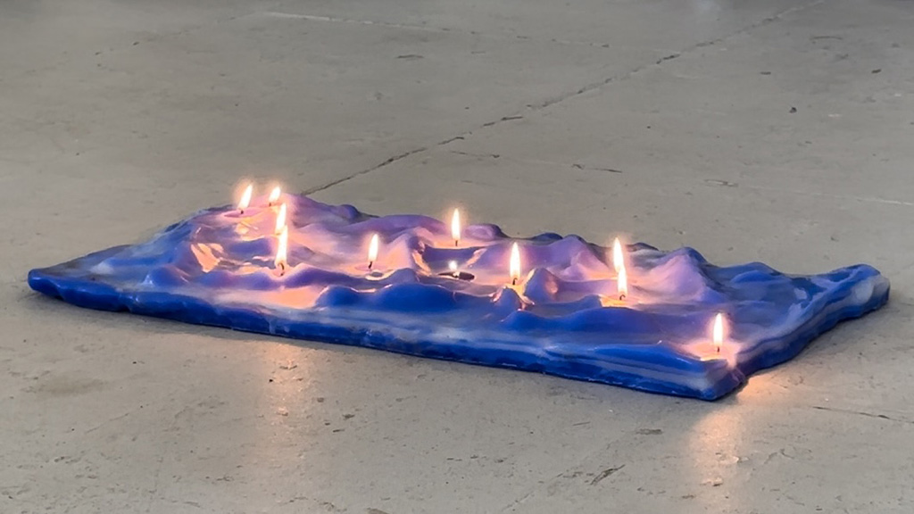 An image of a rectangular block of blue wax holding several burning wicks on a grey floor.