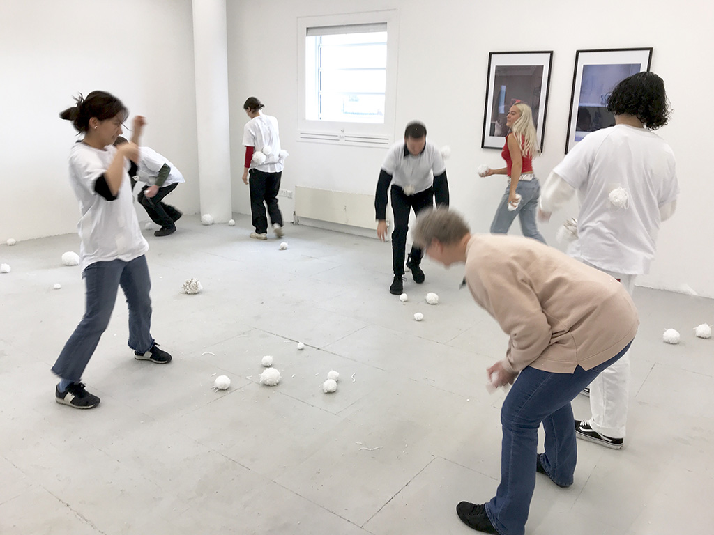 An image of several people participating in a snowball fight with wooden balls, by wearing white, velcro t-shirts, in a grey room.