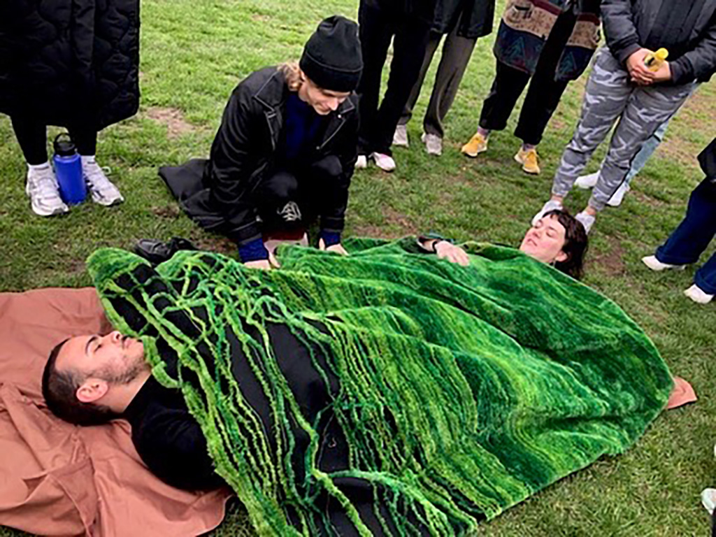 An image of two people under a blanket that is printed to look like grass, on some grass.