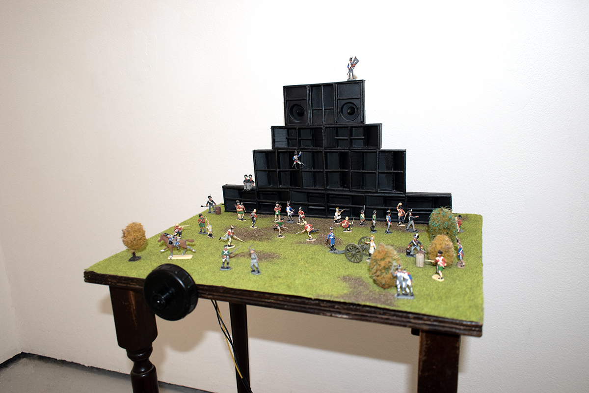 Diorama of a sound system with army figurines. A distance reactive bass tone gets louder as the viewer walks toward the model.