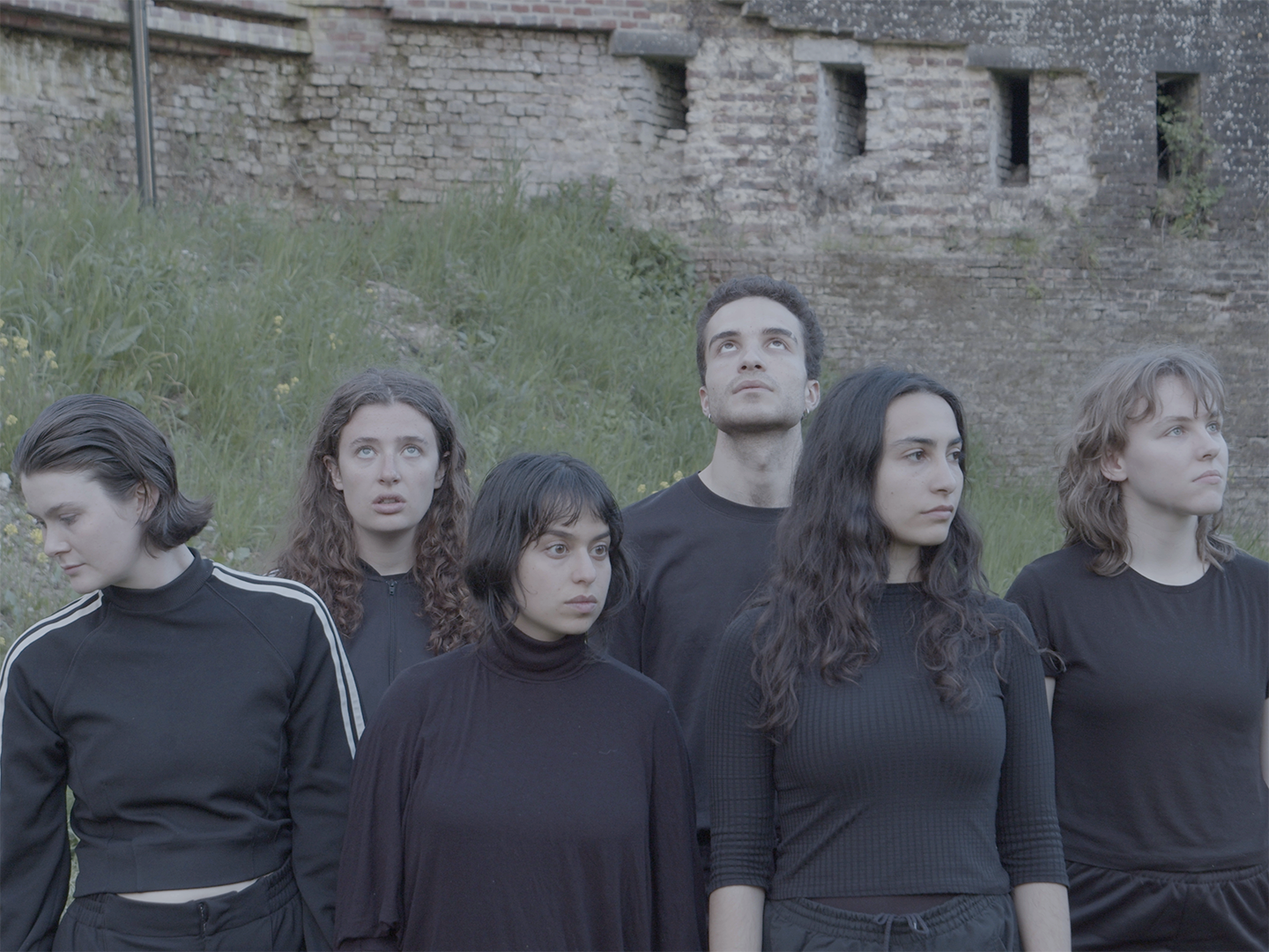 A film still of 6 people dressed in all black, arranged loosely in rows. All people are looking in different directions. The background is a grassy verge in front of a medieval fort built of brick.
