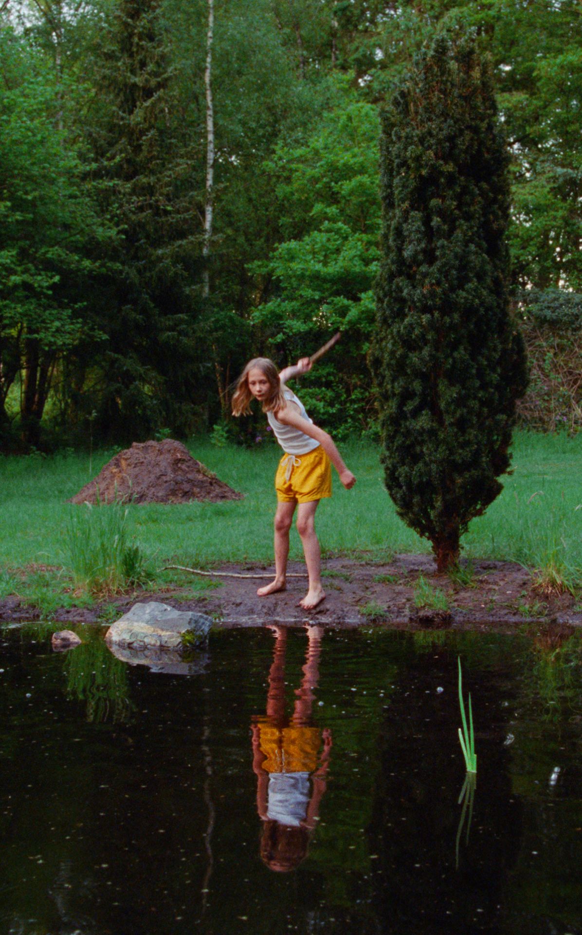 An image of a girl throwing a stick into a lake. Her reflection is in a still, standing pose.
