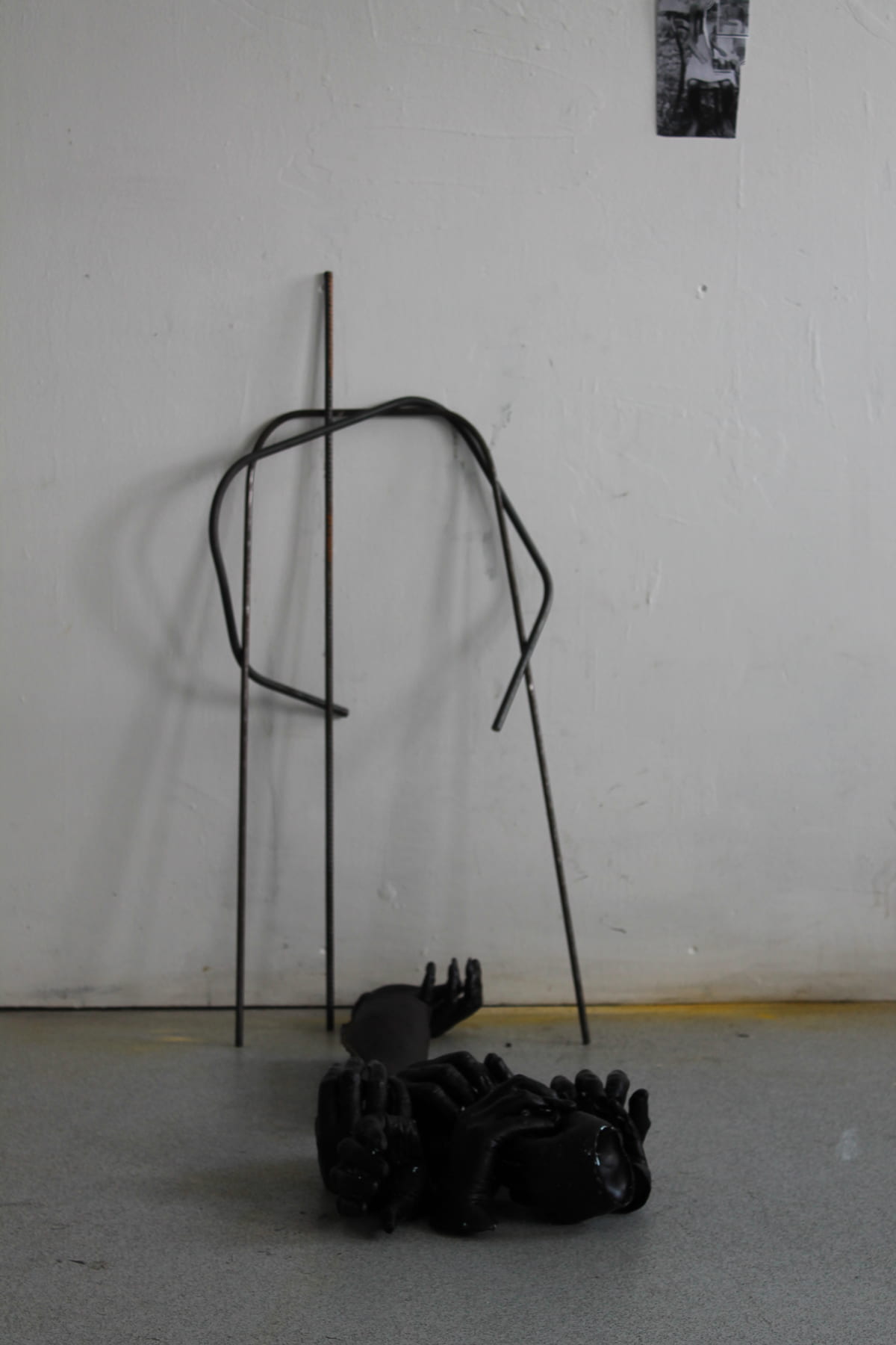 An abstracted figure made of metal rebar is leant against a wall, next to a pile of black, wax-cast hands hands on the floor.