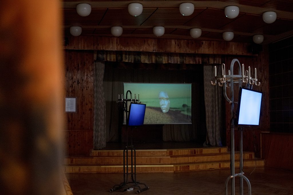 Images of a hall decorated in wooden panneling with a projection screen on the stage and monitors hanging off metal coatrails; image of a flatscreen with yellow image, displayed in a shop window among household goods.
