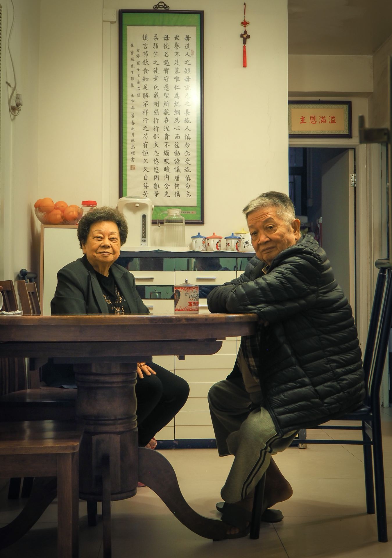 An image of my grandparents facing the camera sitting around a circluar wooden table in their living room in Hong Kong.