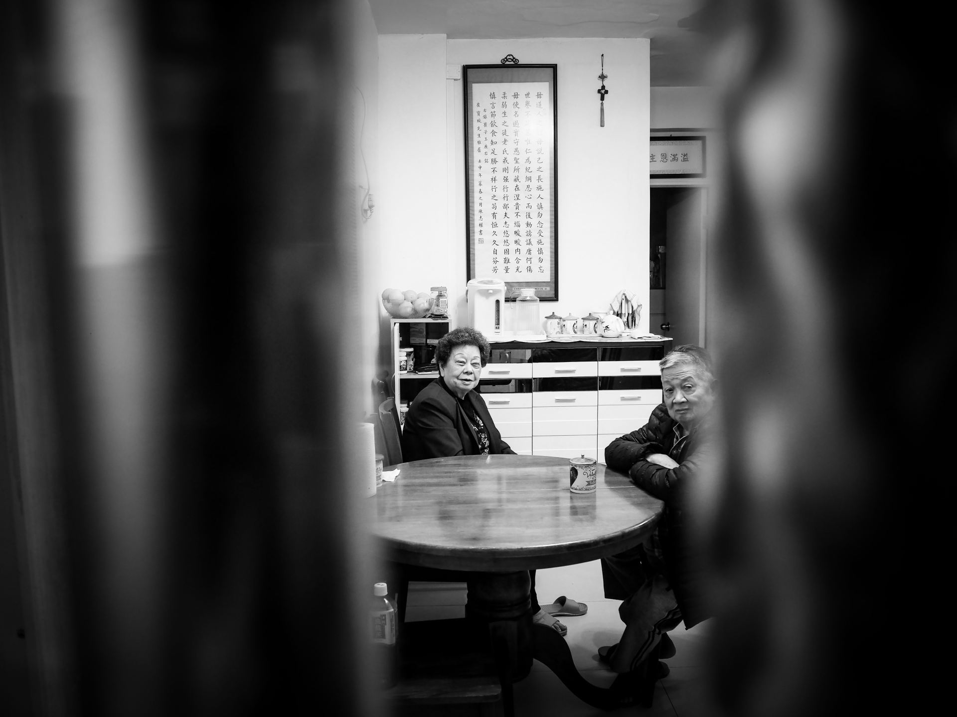 A black and white image of my grandparents facing the camera sitting around a circular wooden table, camera capturing them through a gate.