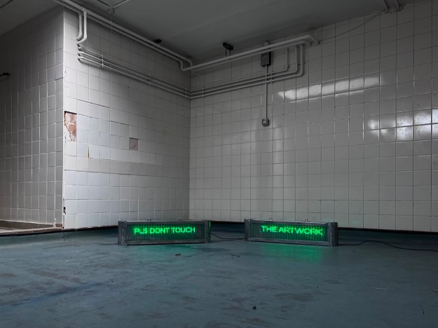 Two LED signs showing green writing on a grey floor in a white, tiled room.