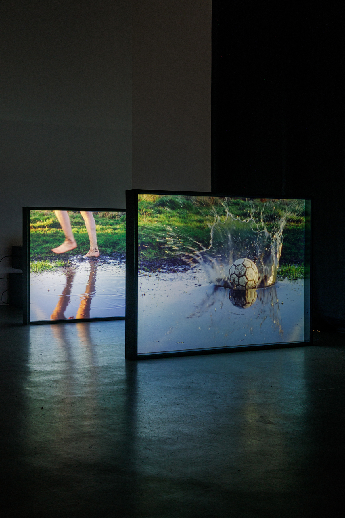 An installation shot of two screens, one depicting a person running through mud, and the other depicting a football in a pond.