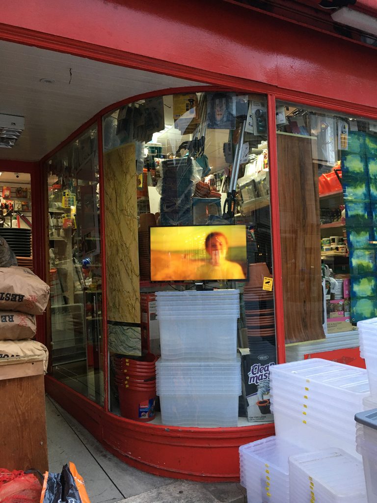 Images of a hall decorated in wooden panneling with a projection screen on the stage and monitors hanging off metal coatrails; image of a flatscreen with yellow image, displayed in a shop window among household goods.

