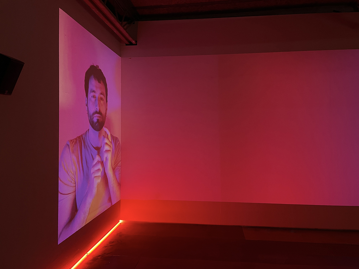 An Image of video projections in a dark room with wooden flooring, depicting a white Mediterenean man making facial expressions in pink background.