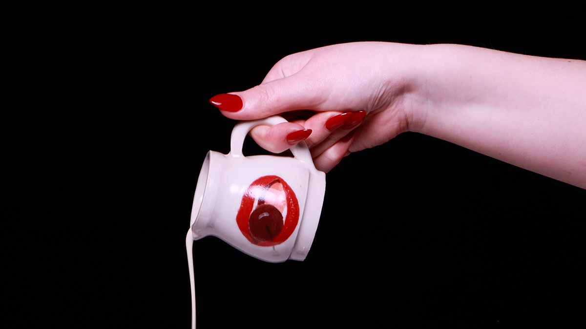 A video still of a hand with red nails pouring milk from a ceramic jug.
