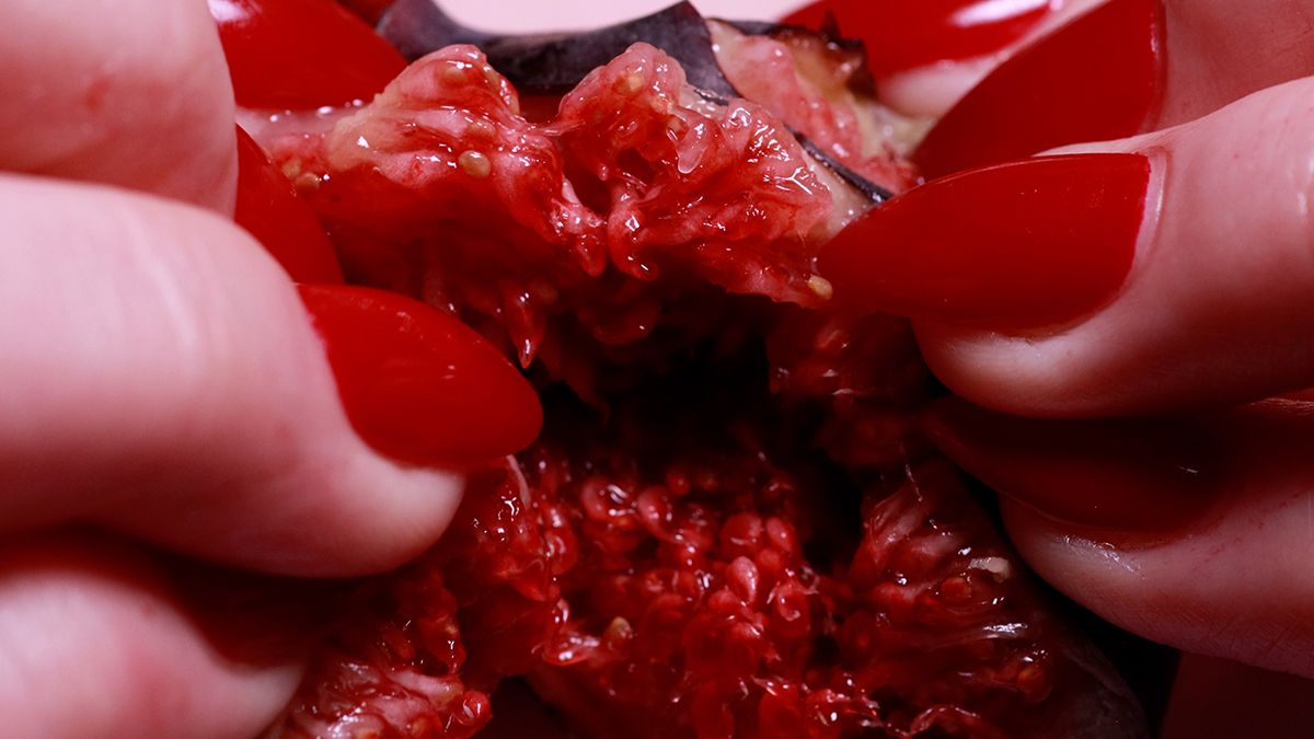 A video still of hands with red nails tearing apart a red fruit.