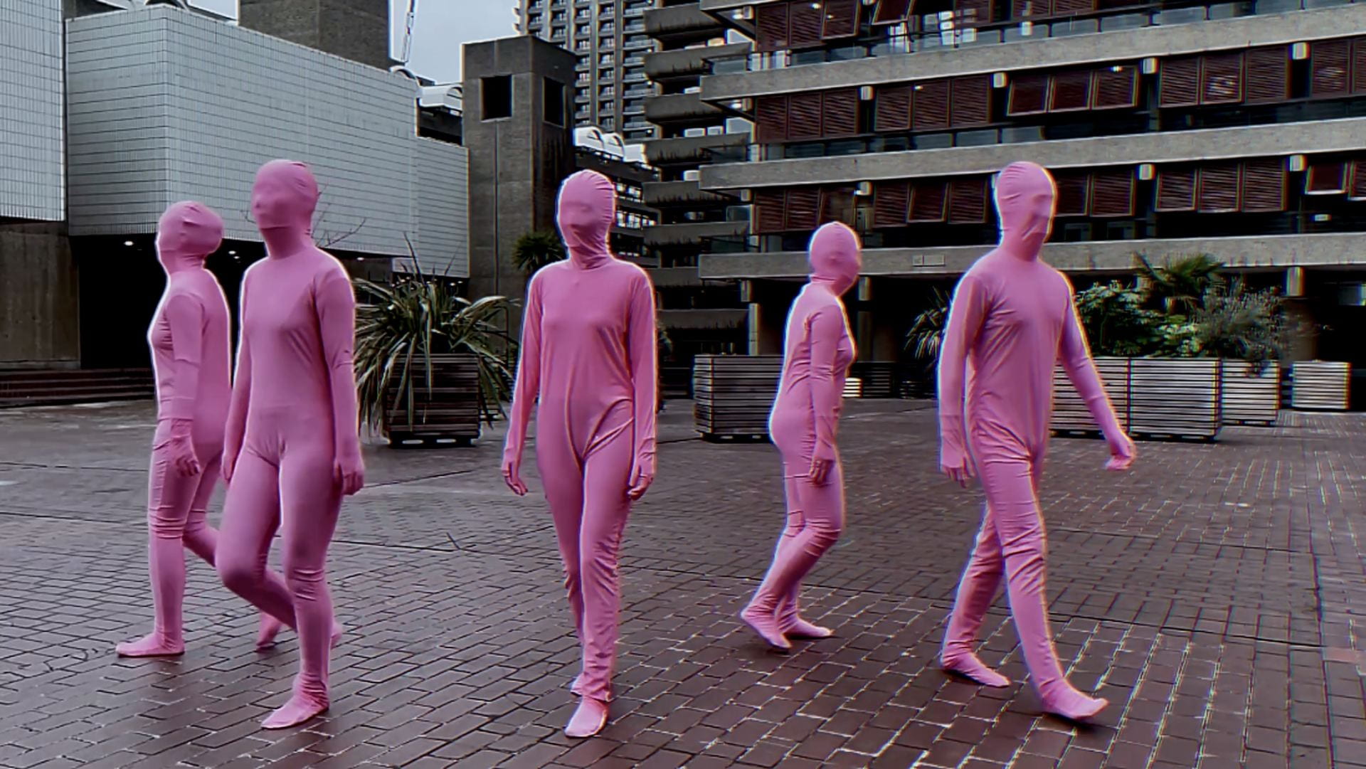 Five people in pink jumpsuits with their faces covered go from moving in unison to moving separately to moving in unison again.
