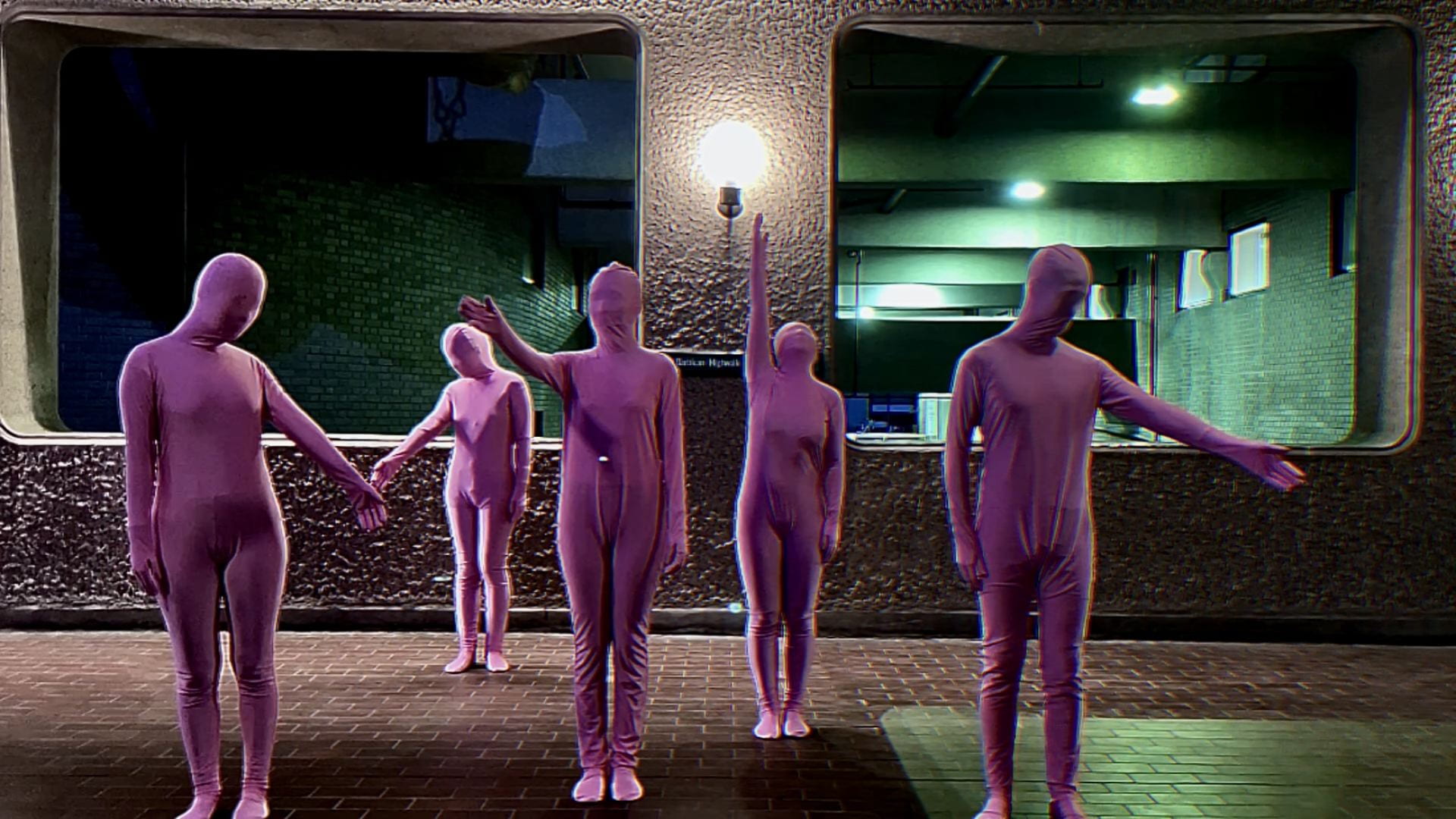 Five people in pink jumpsuits with their faces covered go from moving in unison to moving separately to moving in unison again.