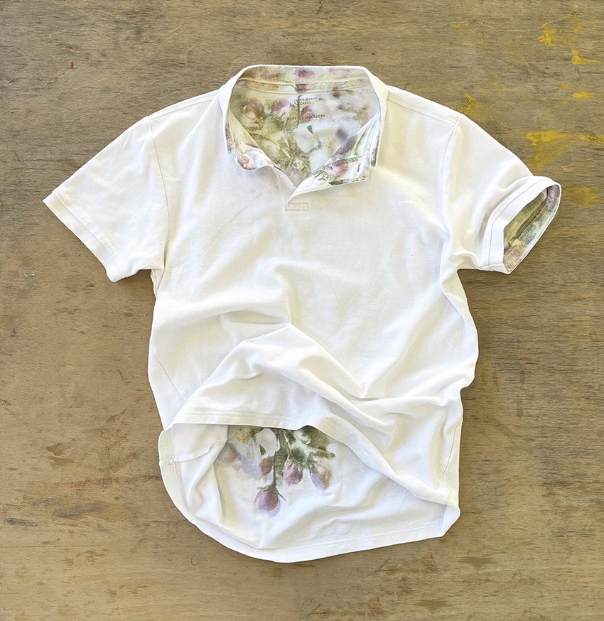 An image of a white t-shirt laying on a wooden floor, printed with purple flowers.