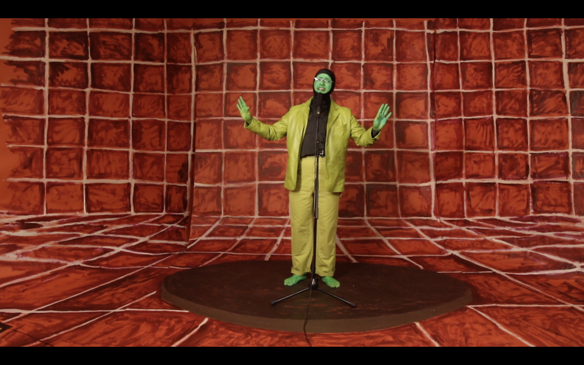 A man in a green suit stands facing a microphone in a painted red tiled space.