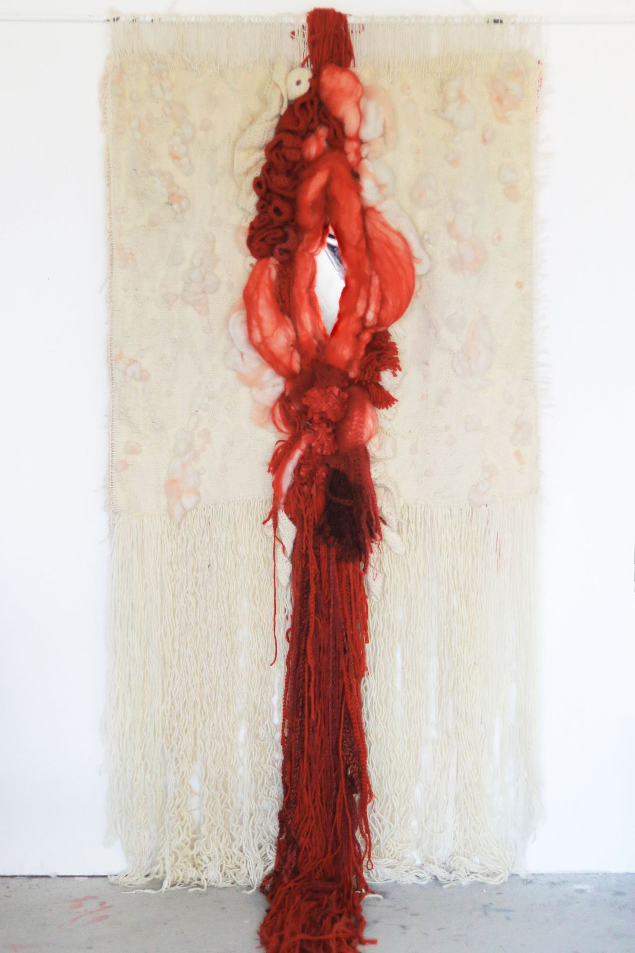 A textile work of an abstracted, red vagina mounted on a cream background and hung on a white wall.
