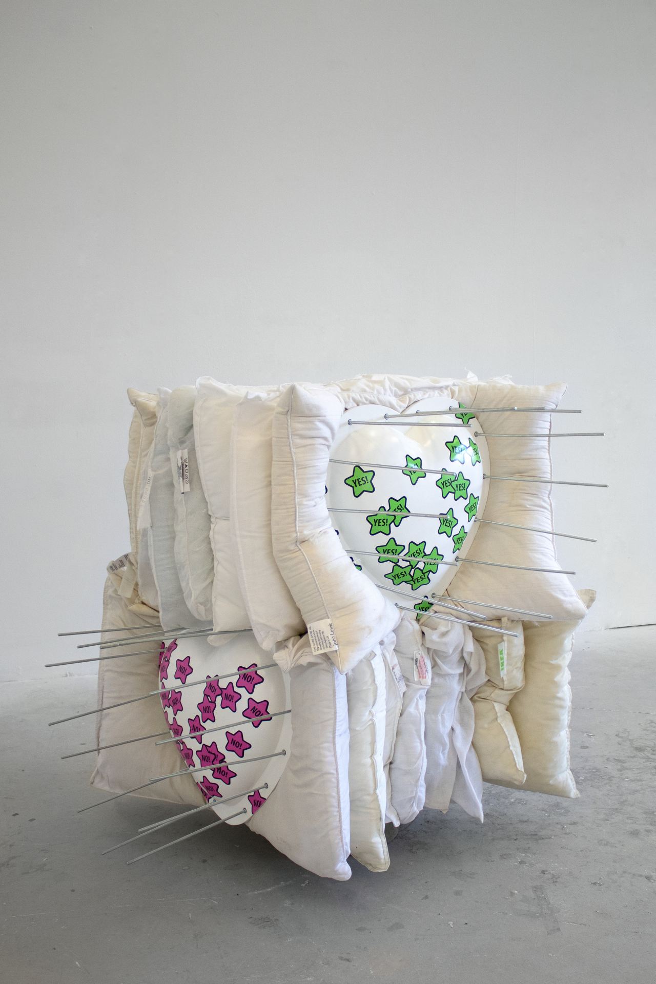 Two piles of pillows stacked one on top of the other and pierced by steel rods between two parts of 3d printed heart shape covered with pink and green star-shaped stickers that read NO! and YES! respectively.