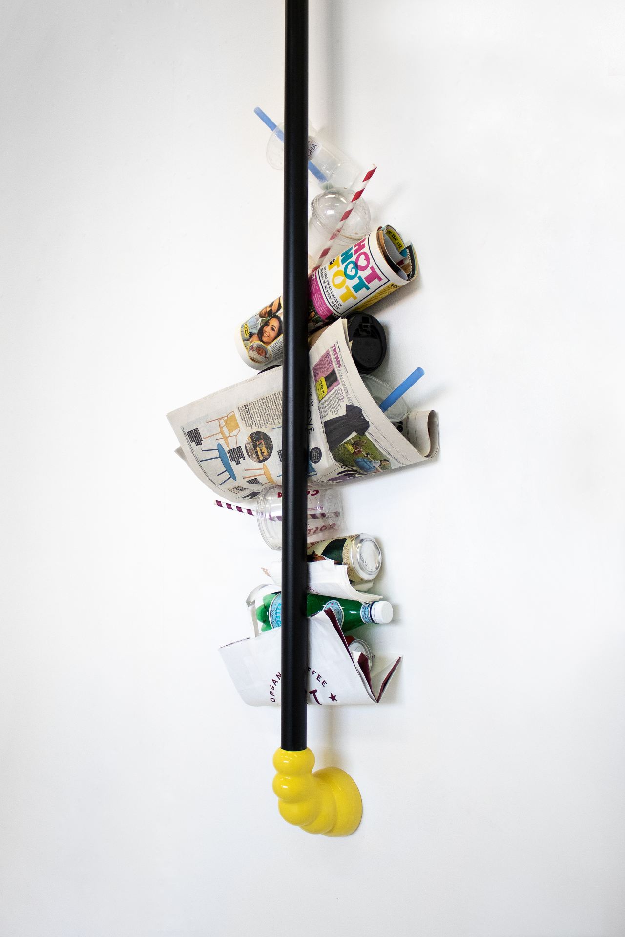Black ABS pipe connected to the wall with two yellow 3d printed fittings. Between the wall to the pipe, there is stacked rubbish; plastic takeaway cups, newspapers and beer cans.  