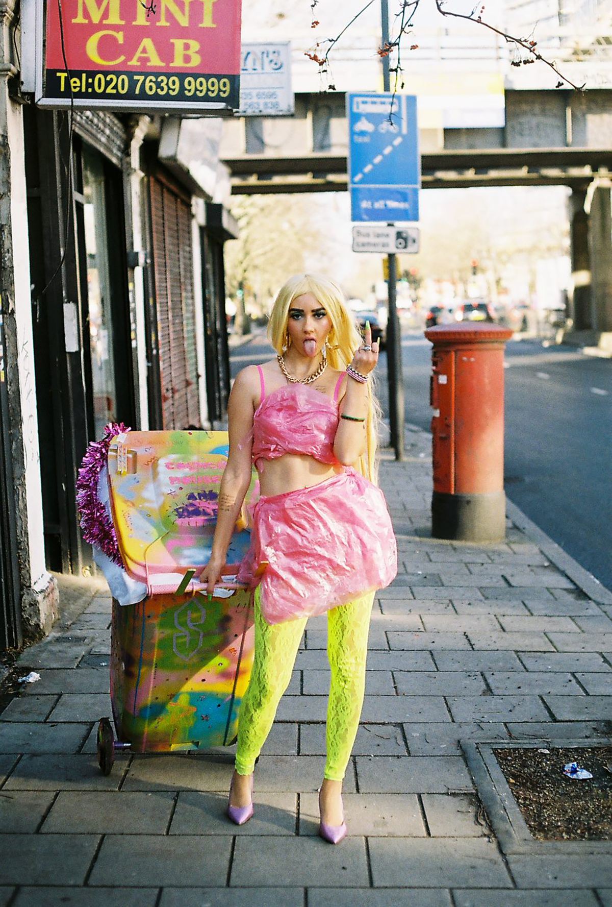 An image of a woman dressed in fluorescent clothes, wheeling a fluorescent wheelie bin down a street, while the other hand shows a middle finger.