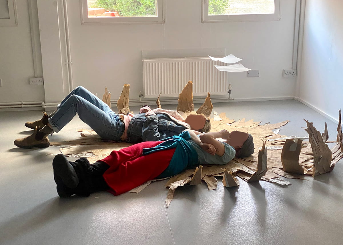 An image of two children lying on a large, floor-based, flat cardboard sculpture.