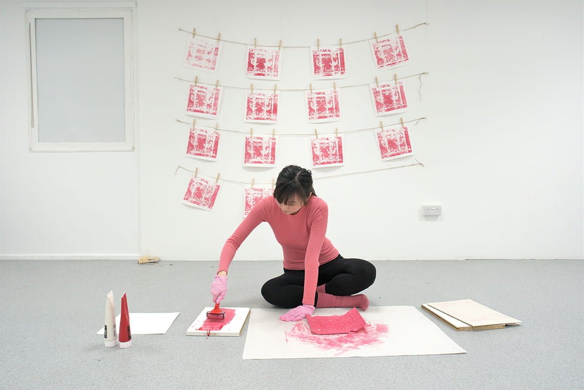 An image of a person sitting on the floor in a pink jumper making prints, some of which are hung up behind her.