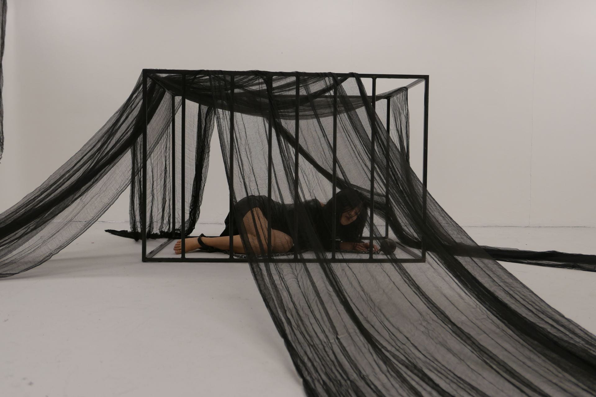 An image of a person dressed in black in a cage in a white room.