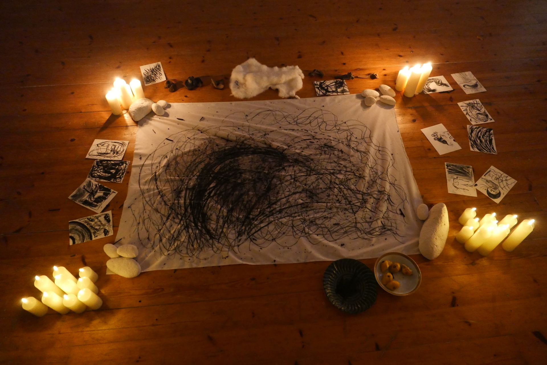 An image of a scribbled drawing on white fabric, surrounded by smaller drawings and candles on a wooden floor.