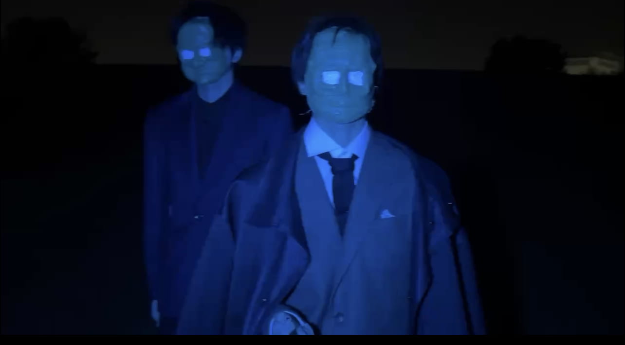 An image of two men with no eyes dressed in suits and steeped in blue light.