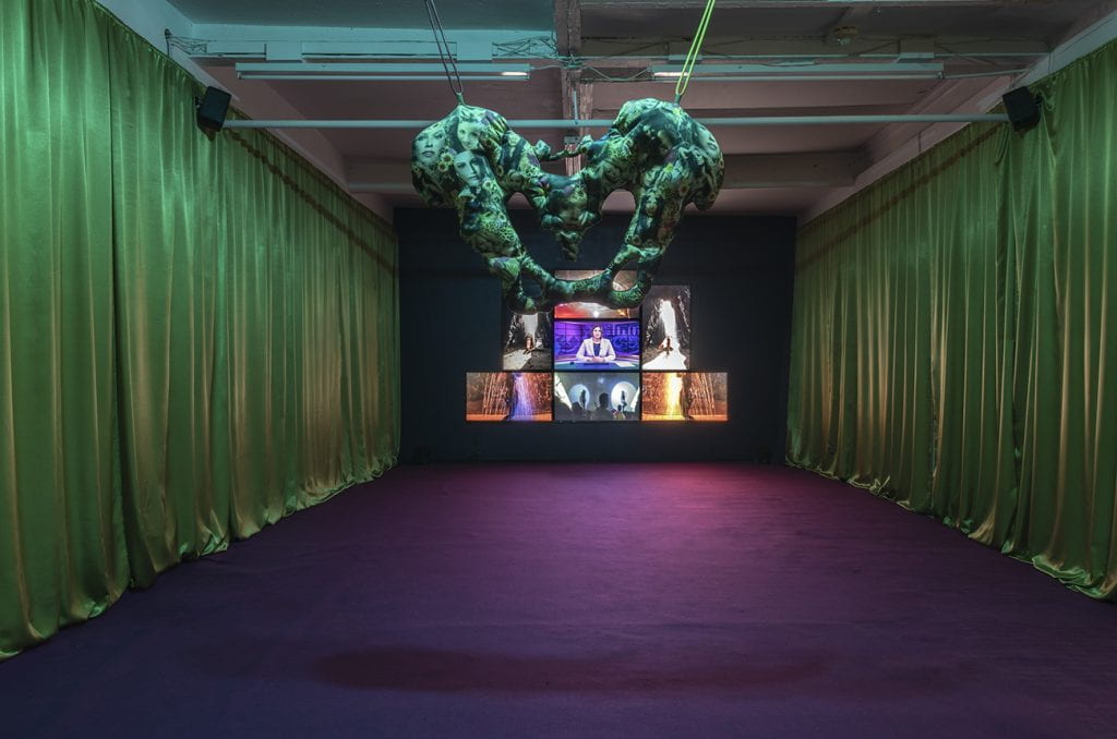 A solo exhibition of Gery Georgieva's work at Cubitt Artists with purple carpet, green curtains and a pyramid of TV screens. 