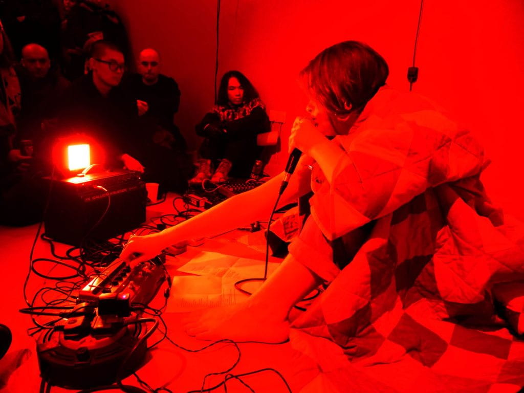 A performance at the opening night of Tiffany Wellington's solo exhibition Grey Area, woman singing into a microphone, sat in a bright red room.