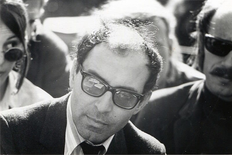 Black and white photographic portrait of Jean-Luc Godard wearing dark glasses at the University of Berkeley in California in 1968.