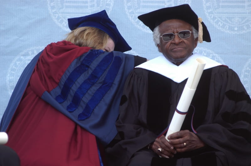 Desmond Tutu at the University of Pennsylvania in 2004 wearing academic gown and mortar board and holding a long white scroll while a woman in academic robes appears to be adjusting something on his back. 
