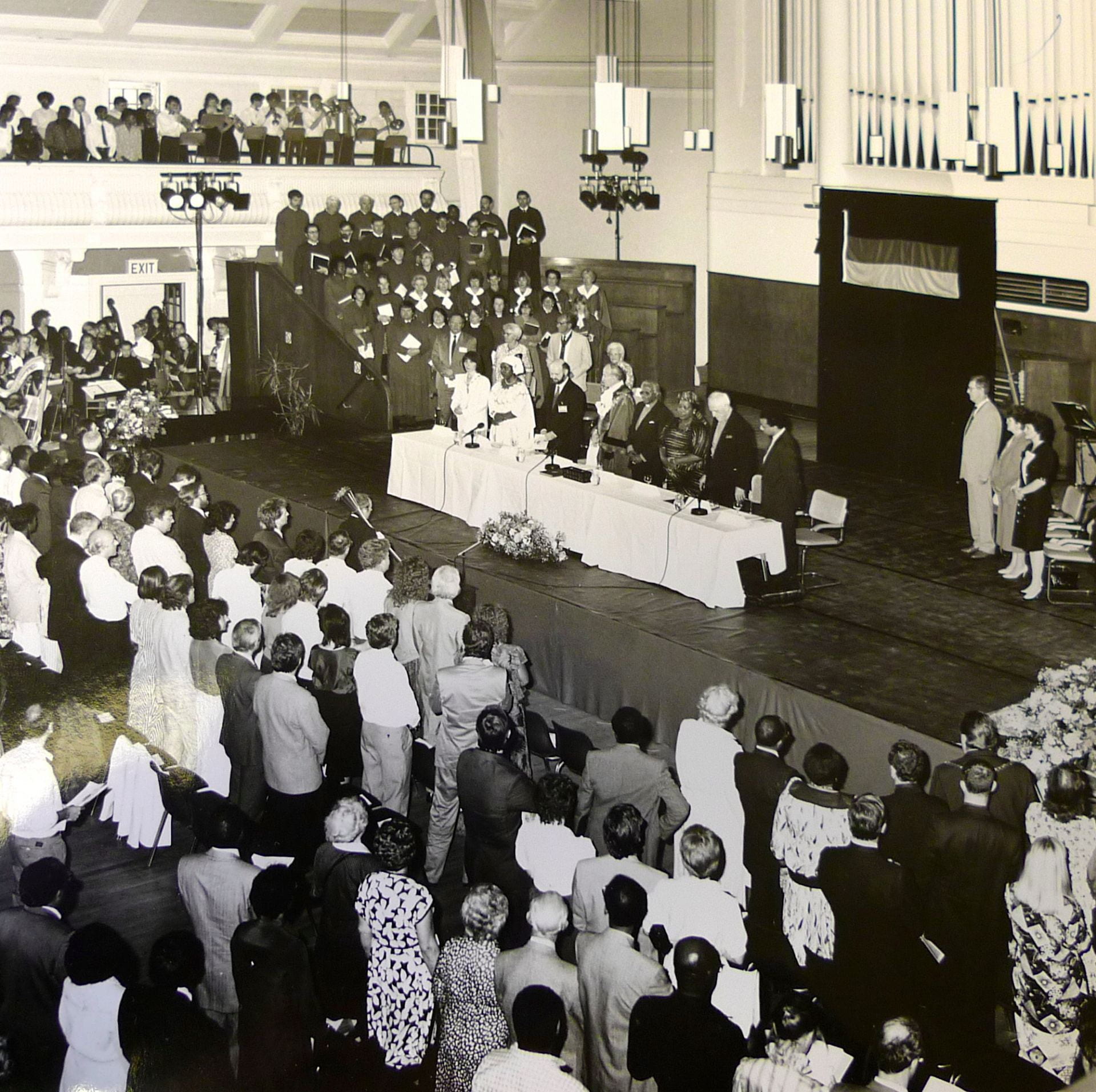 Black and white photograph of the Great Hall of Goldsmiths on May 4th 1990 taken from balcony looking down at stage in front of the organ where his most reverend Desmond Tutu is receiving the honorary freedom of Lewisham. Lewisham Council dignatories, local MP and Goldsmiths' College staff and students present.